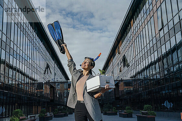 Glückliche Geschäftsfrau mit Taucherflosse und Box vor Bürogebäude
