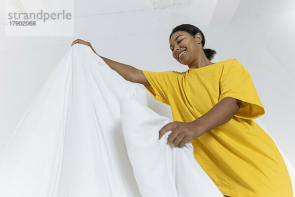 Junge Frau im gelben T-Shirt glättet die Bettdecke im Schlafzimmer