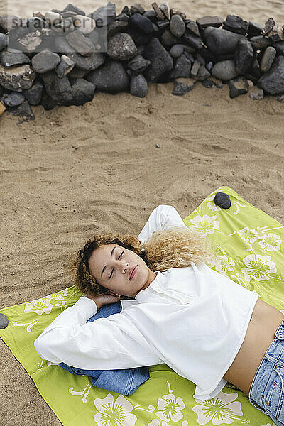 Junge Frau mit Händen hinter dem Kopf schläft auf Picknickdecke am Strand