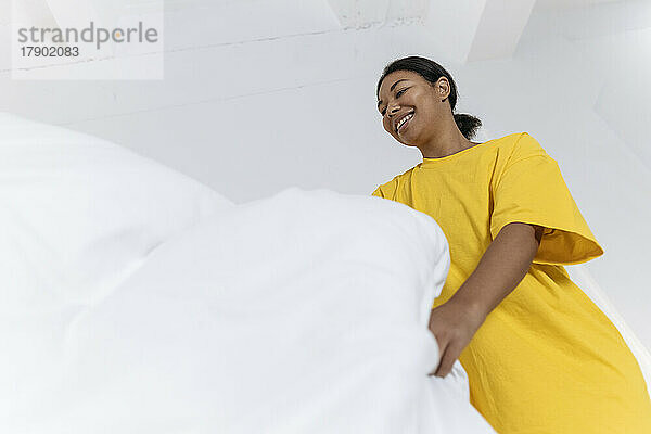 Junge Frau im gelben T-Shirt glättet die Bettdecke im Schlafzimmer