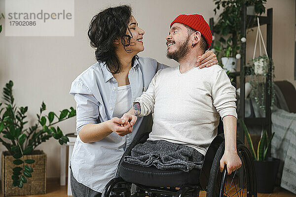 Lächelnder Mann im Rollstuhl hält zu Hause die Hand seiner Freundin