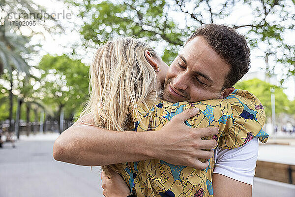 Lächelnder junger Mann mit geschlossenen Augen umarmt Freundin im Park