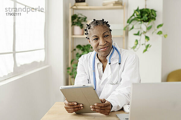 Lächelnde Ärztin mit Tablet-PC sitzt am Tisch im Heimbüro