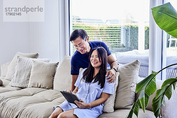 Happy man with woman holding tablet PC sitting on sofa at home