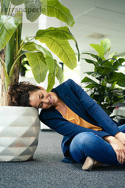 Lächelnde Geschäftsfrau ruht im Büro mit dem Kopf auf einer Topfpflanze