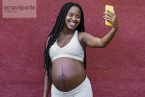 Schwangere Frau macht Selfie mit Smartphone vor der Wand