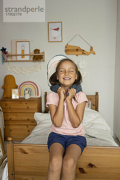 Lächelndes Mädchen sitzt mit geschlossenen Augen und hält einen Spielzeughai im Kinderzimmer