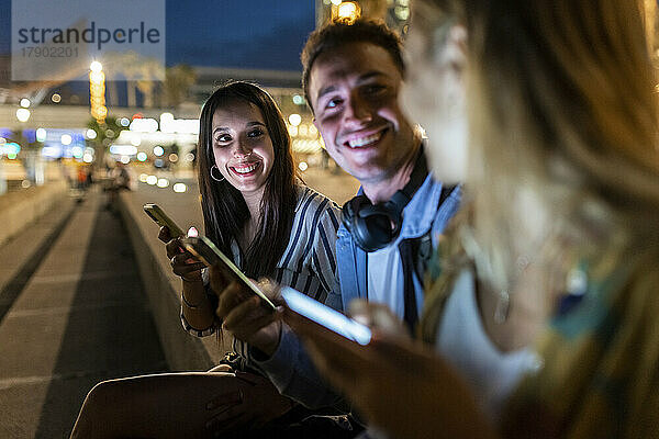 Happy young man and woman holding mobile phones talking with friend at night