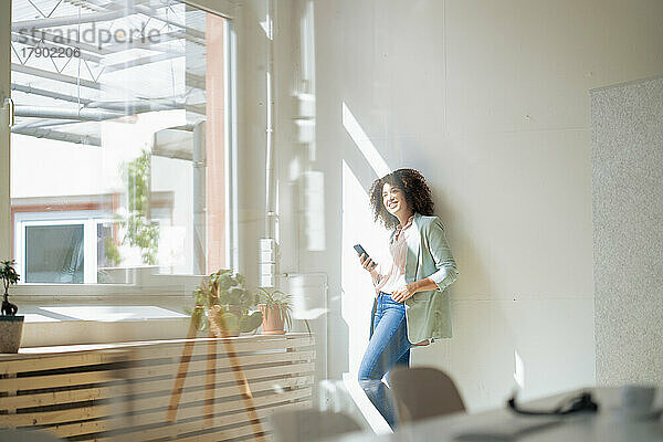 Nachdenkliche Geschäftsfrau mit Smartphone lehnt an der Wand im Büro