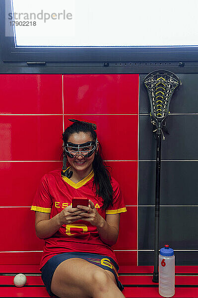 Happy woman wearing protective eyewear sitting with mobile phone in locker room