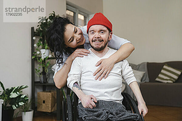 Lächelnde Frau umarmt ihren Freund im Rollstuhl zu Hause