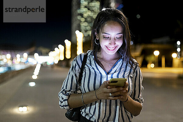 Lächelnde junge Frau  die nachts Textnachrichten über das Mobiltelefon sendet