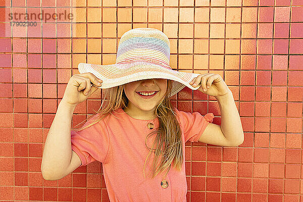 Smiling girl covering face with hat in front of wall