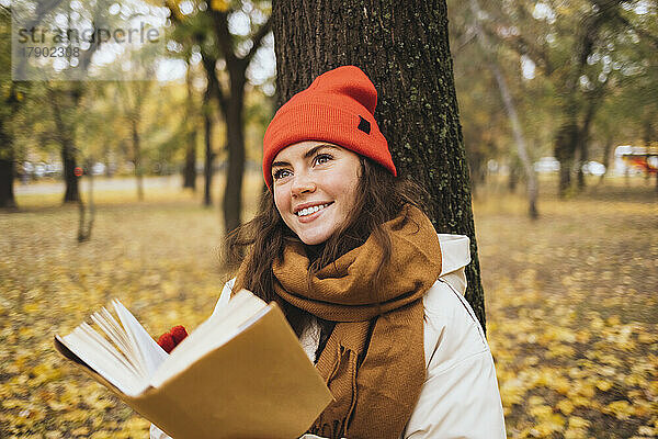 Nachdenklich lächelnde junge Frau mit Buch vor Baumstamm im Park