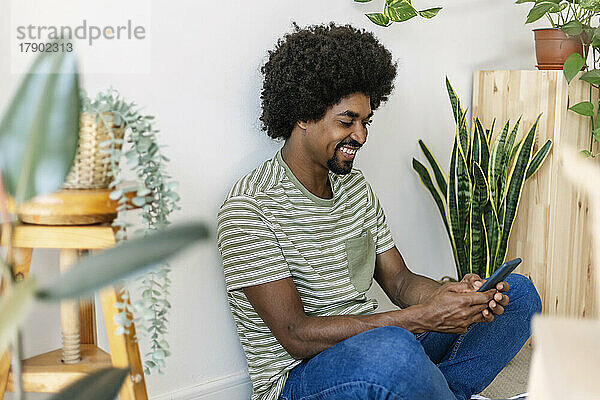 Glücklicher Mann mit Afro-Frisur  der zu Hause sein Smartphone nutzt