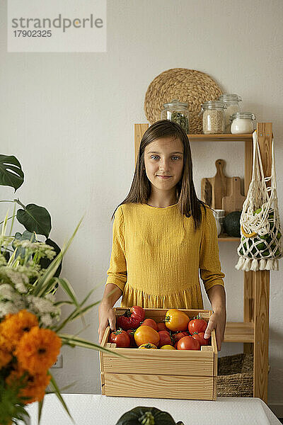 Smiling girl holding crate of tomatoes by table at home
