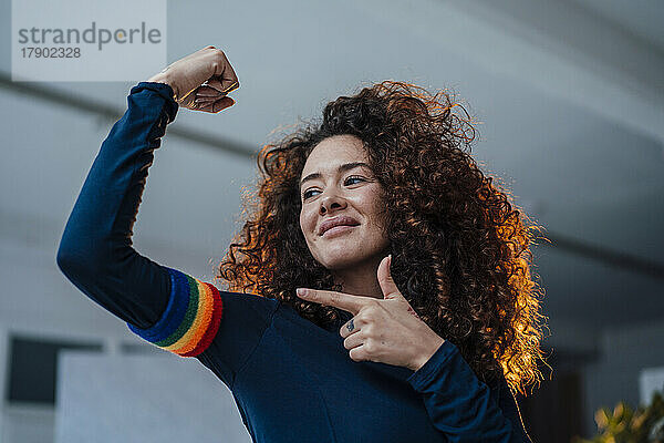 Lächelnde junge Frau  die ihre Muskeln spielen lässt und auf das Regenbogenarmband zeigt