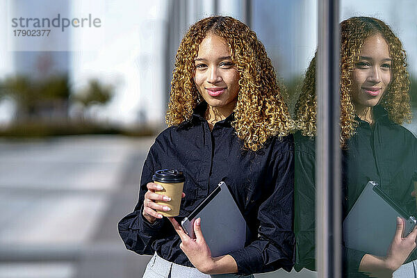 Lächelnde junge Geschäftsfrau hält Kaffeetasse in der Hand und stützt sich auf Glas