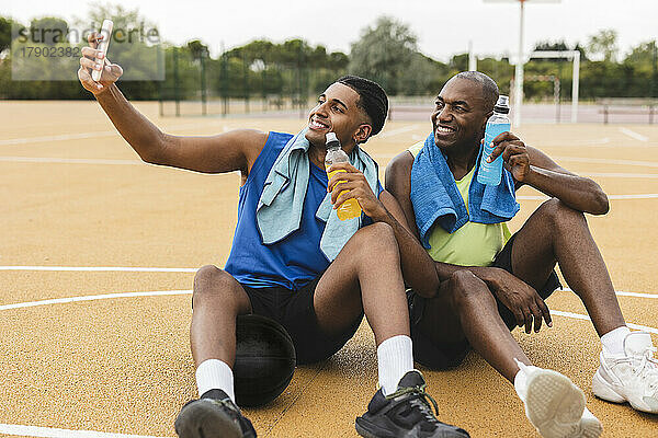 Sohn macht Selfie per Smartphone und sitzt mit Vater auf dem Basketballplatz