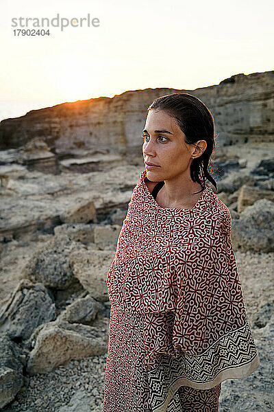 Thoughtful woman wrapped in blanket standing in front of rock formation
