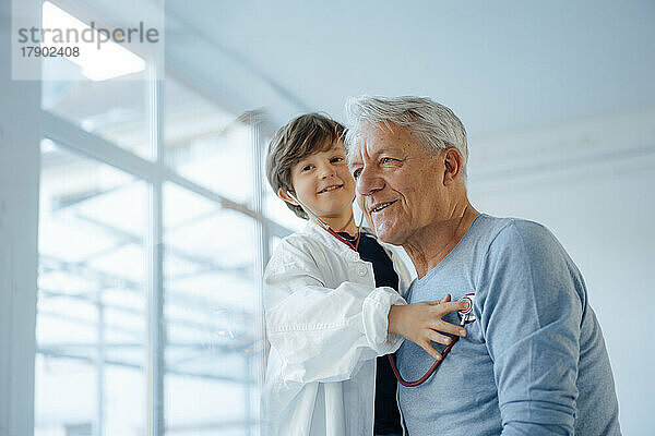 Lächelnder Junge  der es als Arzt nachahmt  der zu Hause Großvater mit Stethoskop überprüft