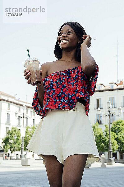 Fröhliche junge Frau mit der Hand im Haar und einer Einweg-Kaffeetasse