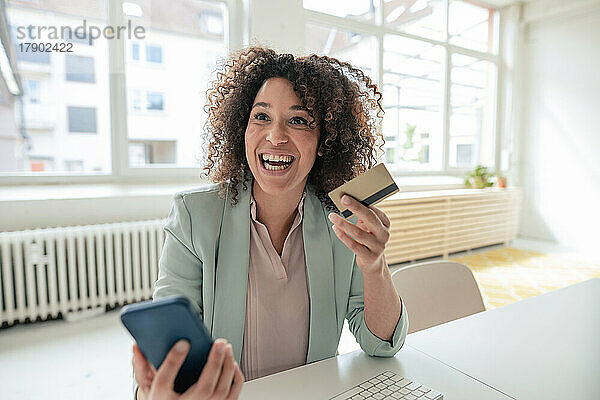 Fröhliche Geschäftsfrau mit Kreditkarte und Smartphone am Arbeitsplatz