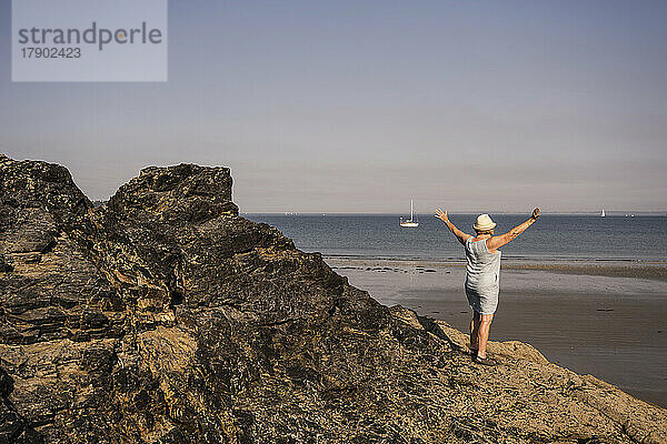 Ältere Frau mit ausgestreckten Armen steht auf einem Felsen am Strand