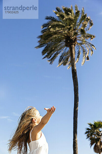 Mädchen mit ausgestrecktem Arm steht an einem sonnigen Tag vor einer Palme