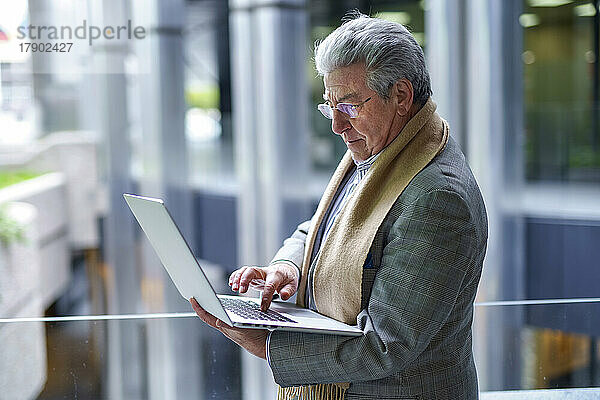 Leitender Geschäftsmann arbeitet am Laptop und steht am Glasgeländer