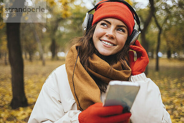 Lächelnde junge Frau hält ihr Mobiltelefon in der Hand und hört im Park Musik über Kopfhörer