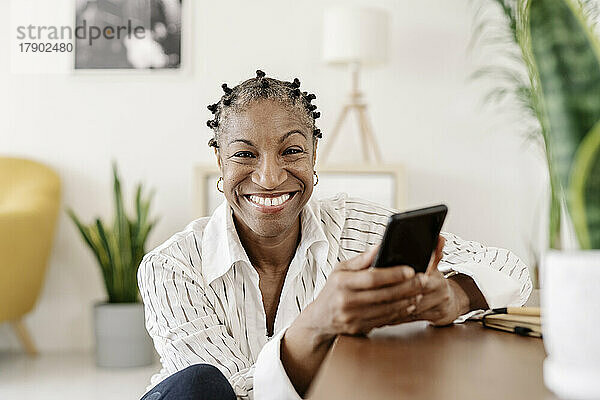 Glückliche reife Frau mit Smartphone sitzt zu Hause im Wohnzimmer