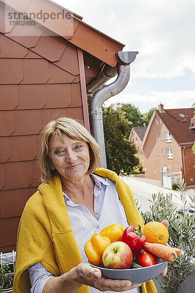 Lächelnde blonde Frau mit einer Schüssel frischem Obst und Gemüse auf dem Balkon