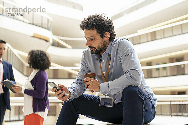 Geschäftsmann schreibt Textnachrichten auf dem Smartphone mit Kollegen  die im Hintergrund im Büro diskutieren