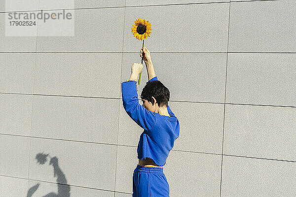 Frau mit erhobenen Armen hält an einem sonnigen Tag eine Sonnenblume an der Wand