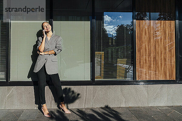 Lächelnde Geschäftsfrau  die an einem sonnigen Tag vor einer Glaswand mit ihrem Smartphone spricht