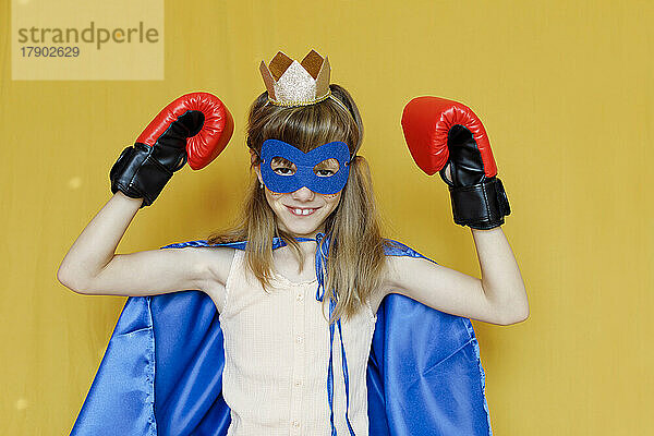 Girl in cape wearing mask and boxing gloves against yellow background