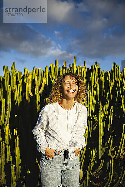 Glückliche junge Frau mit den Händen in den Taschen  die vor einem Kaktus steht