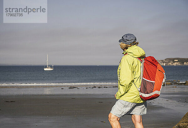 Ältere Frau mit Rucksack steht an einem sonnigen Tag am Strand