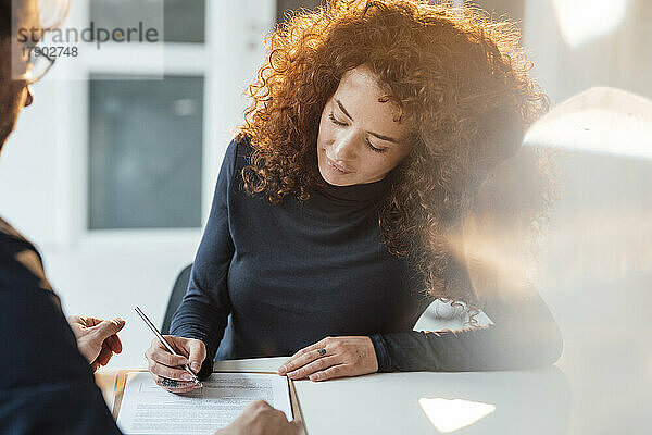 Geschäftsfrau mit lockigem Haar unterschreibt Dokument am Schreibtisch im Büro