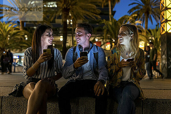 Fröhliche Freunde halten nachts Smartphones in der Hand und reden miteinander