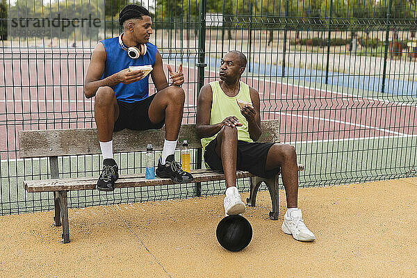 Vater und Sohn essen Sandwiches und sitzen auf einer Bank am Basketballplatz