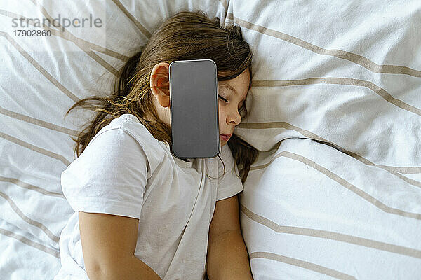 Girl with smart phone on cheek sleeping on bed at home