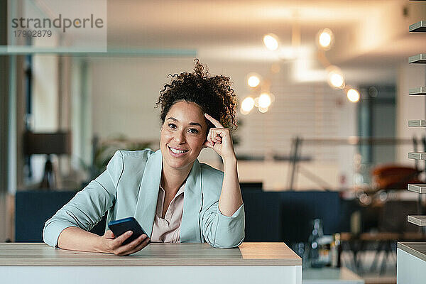 Lächelnde Geschäftsfrau mit Mobiltelefon am Schreibtisch im Büro