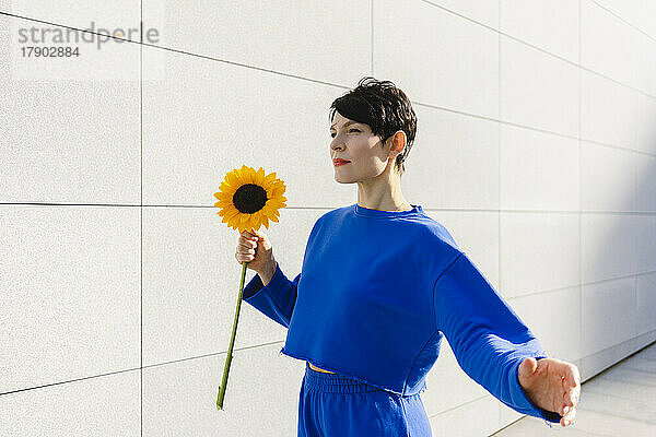 Frau mit kurzen Haaren hält an einem sonnigen Tag eine Sonnenblume an der Wand