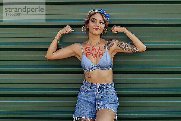 Frau mit Girl-Power-Text auf der Brust  der Bizeps vor einer grünen Metallwand zeigt