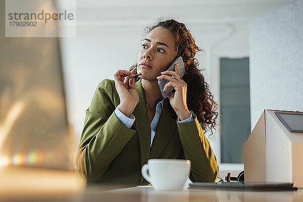 Nachdenkliche Geschäftsfrau  die am Schreibtisch im Büro neben einem Musterhaus sitzt und auf einem Smartphone spricht