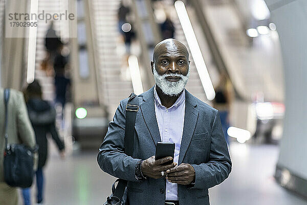 Lächelnder älterer Geschäftsmann mit Laptoptasche und Mobiltelefon am Bahnhof