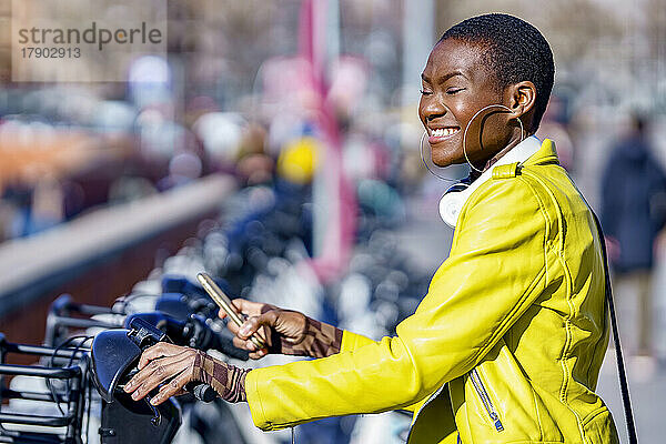Glückliche Frau in Jacke  die an einem sonnigen Tag an der Fahrradstation steht