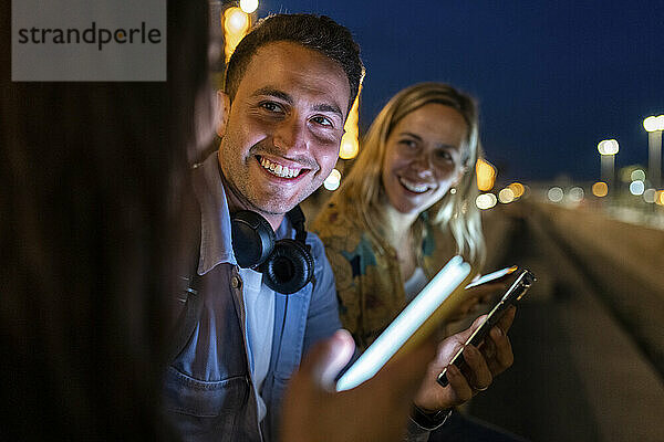 Happy young man with smart phone sitting amidst friends at night